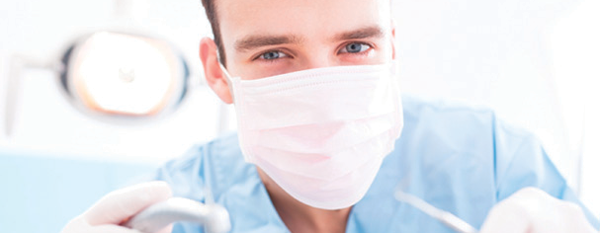 A dentist with a mask, holding dental implements, and looking at the camera.