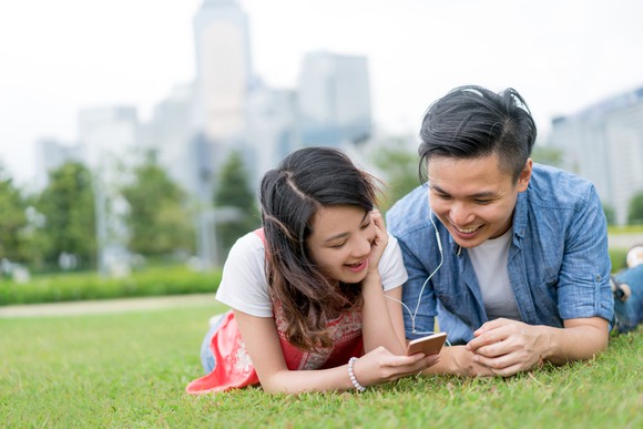 A man and woman lie on the grass with earplugs in their ears, looking at a smartphone.