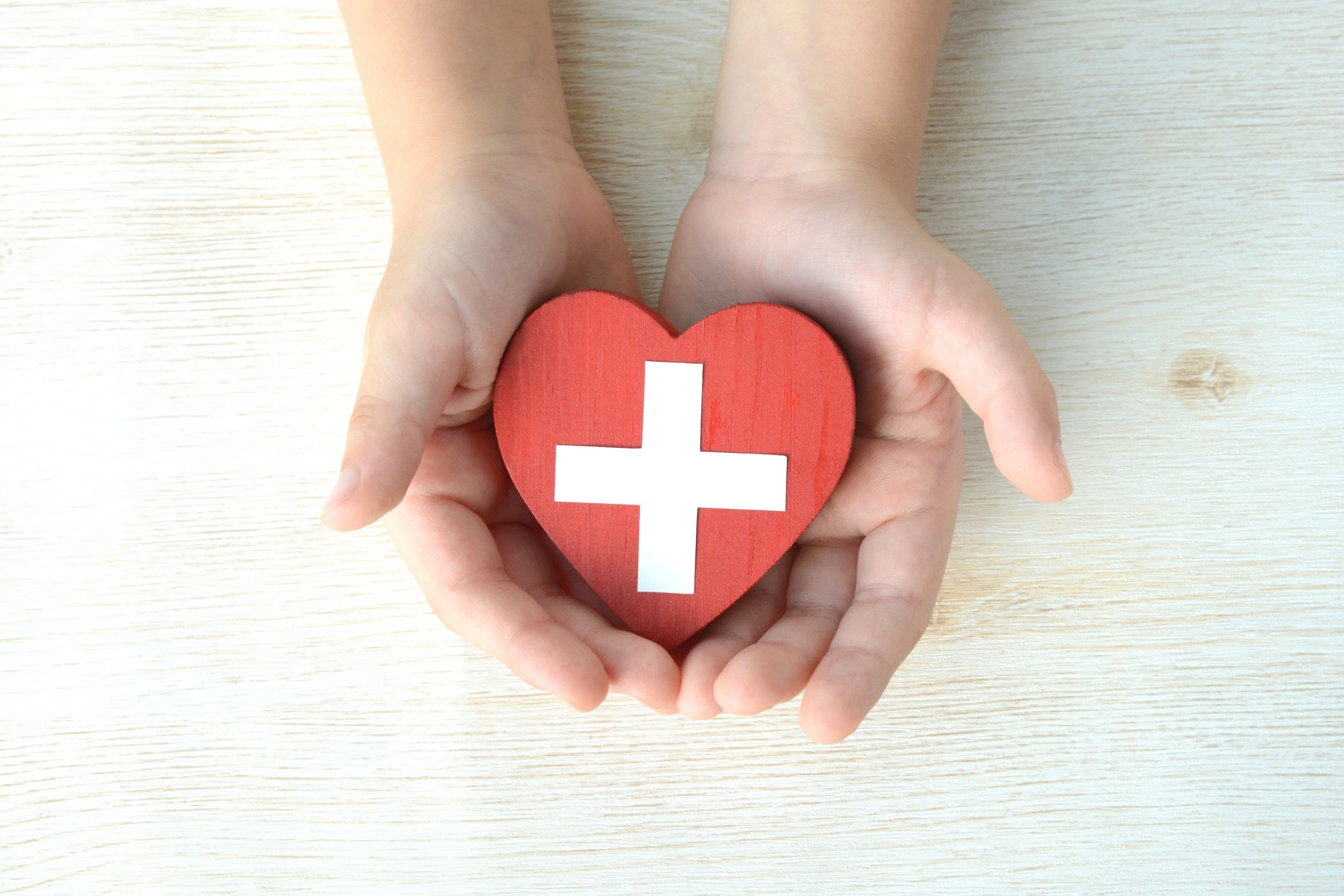 Two outstretched hands hold a small plastic heart toy with a white cross medical symbol painted on it.