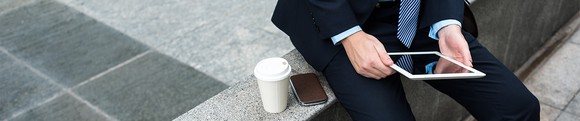 A seated man in a suit, holding a tablet, with a smartphone and a cup of coffee next to him.