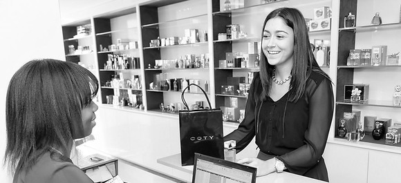 A woman behind a counter hands a makeup bag to another woman in a black and white photo. 