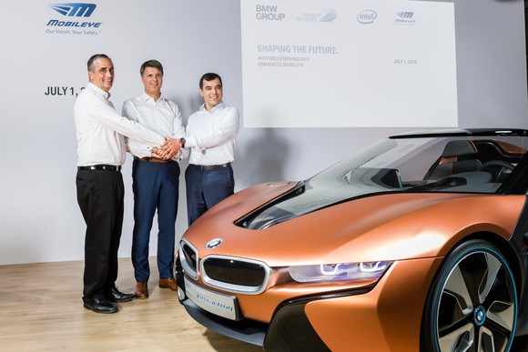 The three are shown on a stage with an orange BMW i8 sports car. 