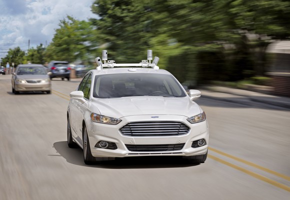 A white Ford Fusion sedan with self-driving sensor hardware on a suburban road