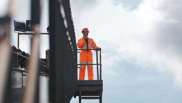 A worker in an orange jumpsuit standing on a catwalk talking into a mobile phone.