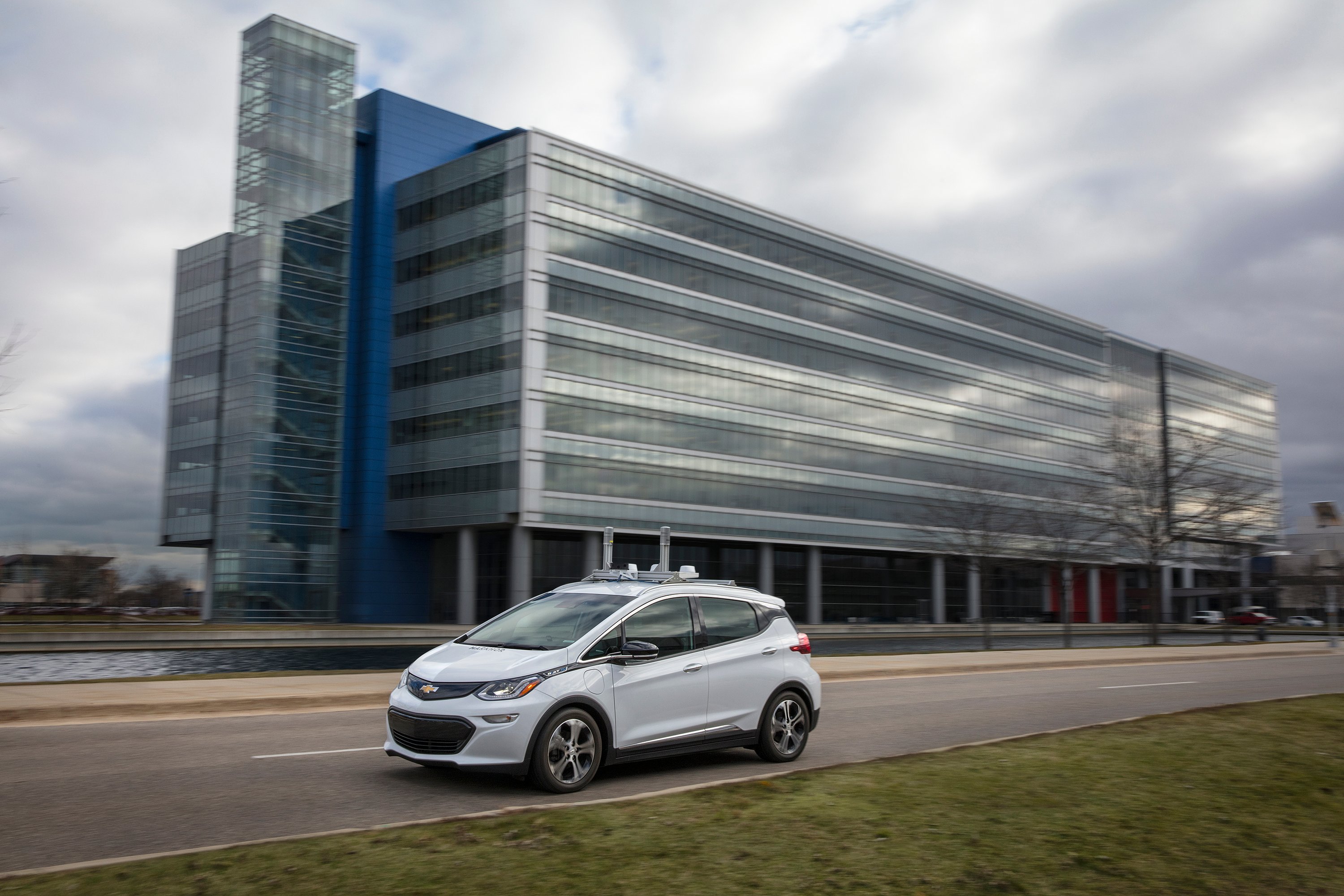 A white Chevrolet Bolt EV with self-driving sensors is shown on a public road near GM's technical center.