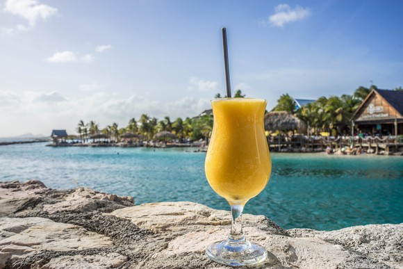 A fruity drink is positioned on a rock in a tropical setting.