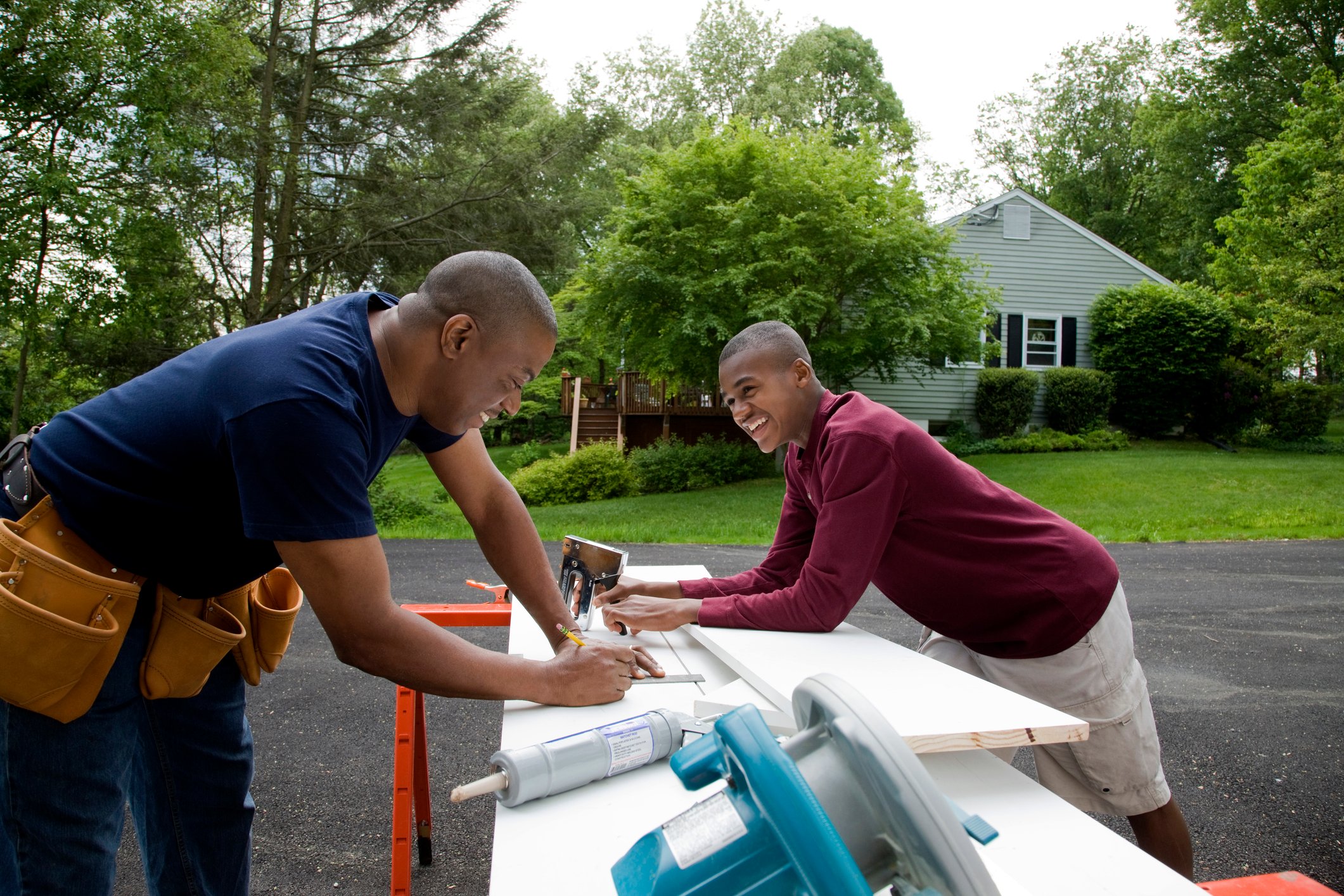 Two smiling men with tools and tool belts work outdoors on a project.