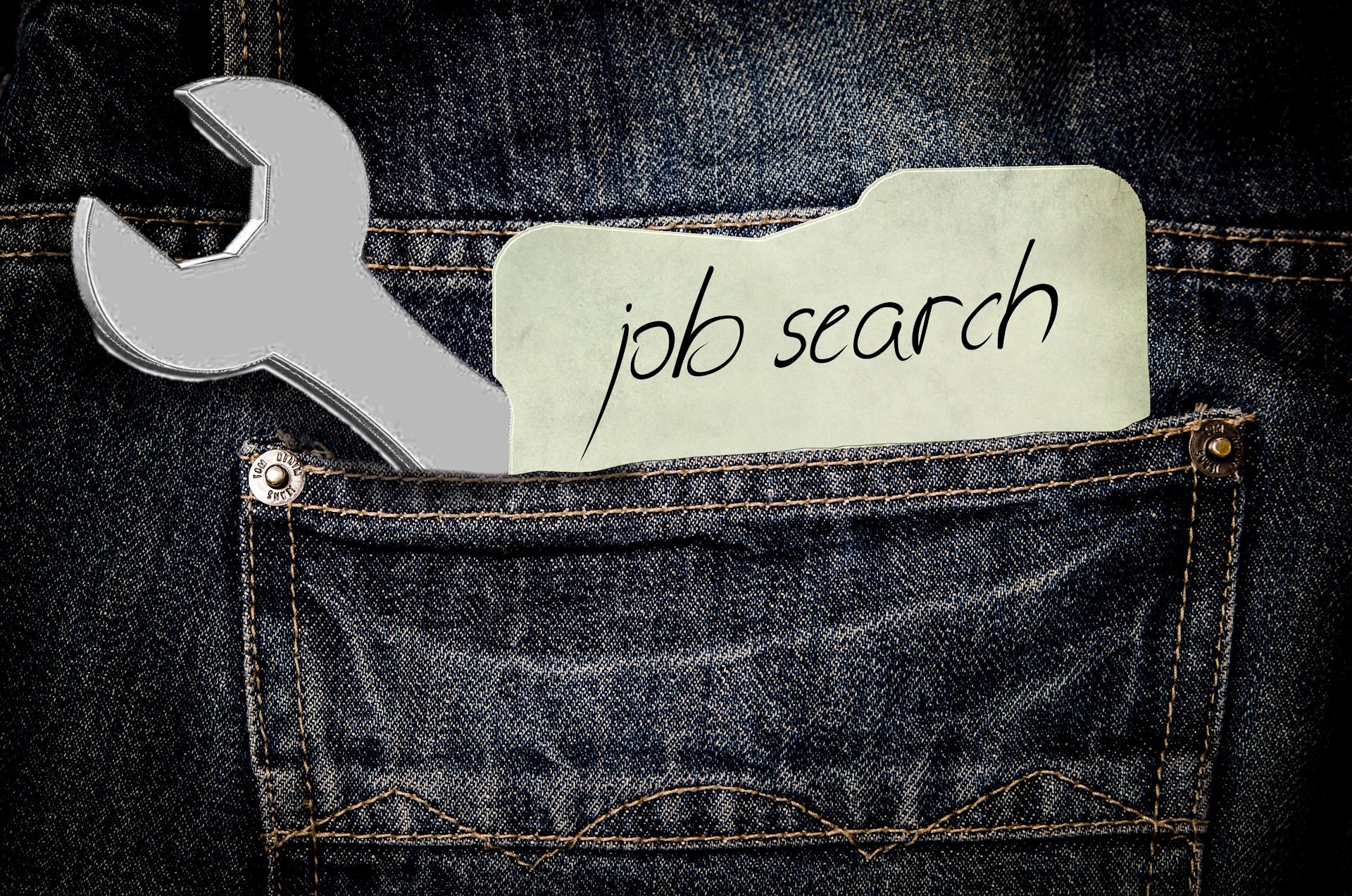 A wrench and a note that says "job search" are sticking out of the pocket of a pair of jeans.