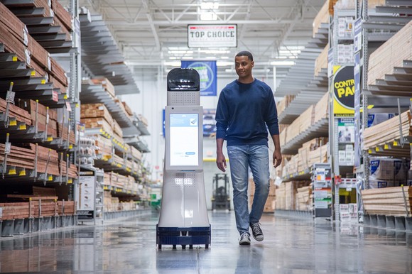 A LoweBot with a shopper walking alongside it. LoweBots look like a rolling aisle display, but have a touch screen on their front for shoppers to interact with them.