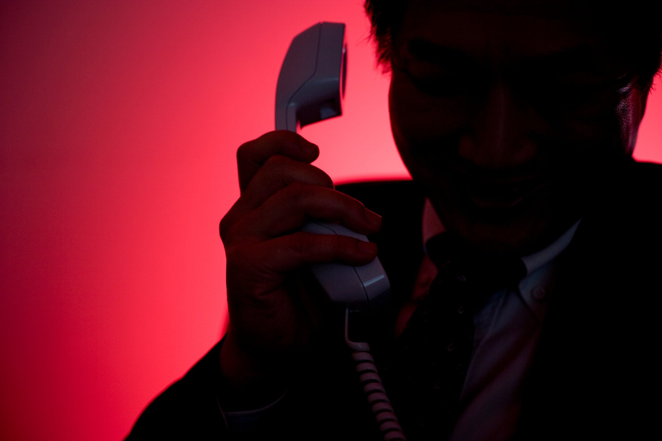 A man silhouetted against a red background raises a phone to his ear. 