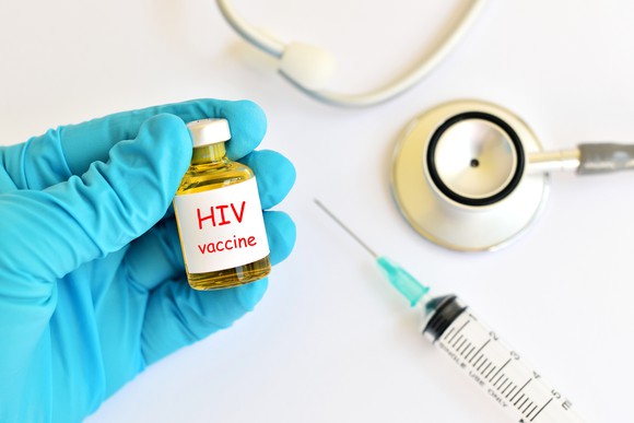 Hand holding bottle that says HIV vaccine, with a syringe and a stethoscope next to it.