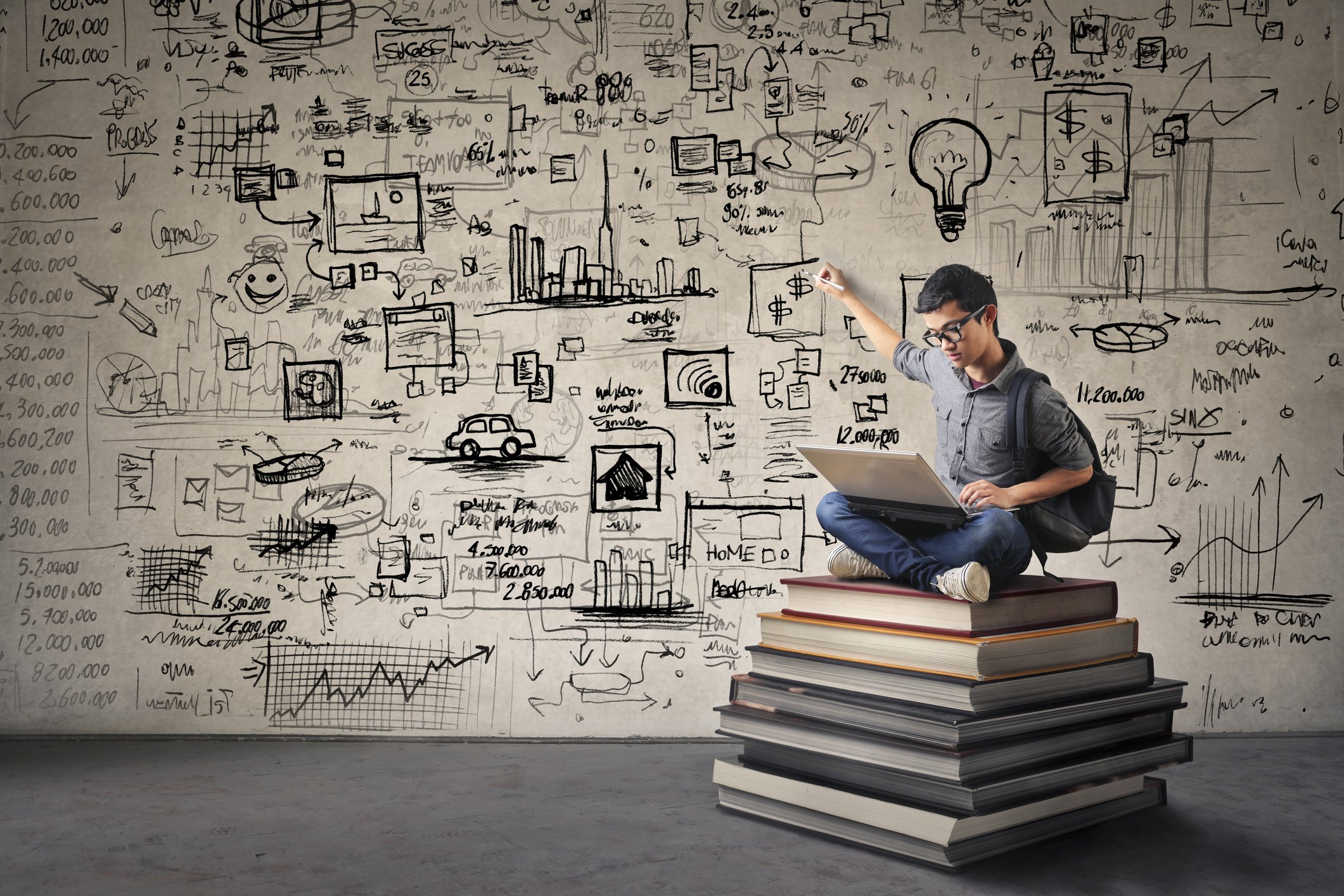 A man, using a laptop, sits cross-legged on a pile of oversized books, as he points to a cluster of dollar signs on the wall behind him.