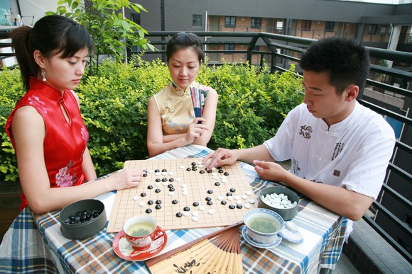 Three people sitting at a table while two play the Chinese game of Go.