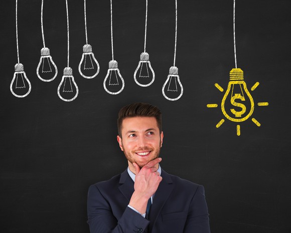A man, standing under an illustration of a cluster of unlit light bulbs, smiles at a bulb illuminating a dollar sign.