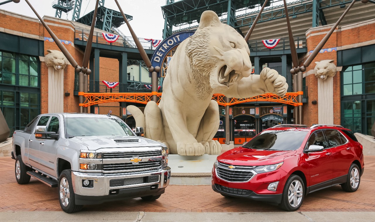 A Silver Ice Metallic 2018 Silverado 2500HD and a Cajun Red 2018 Equinox, parked side by side under a tiger statue at Comerica Park in Detroit.
