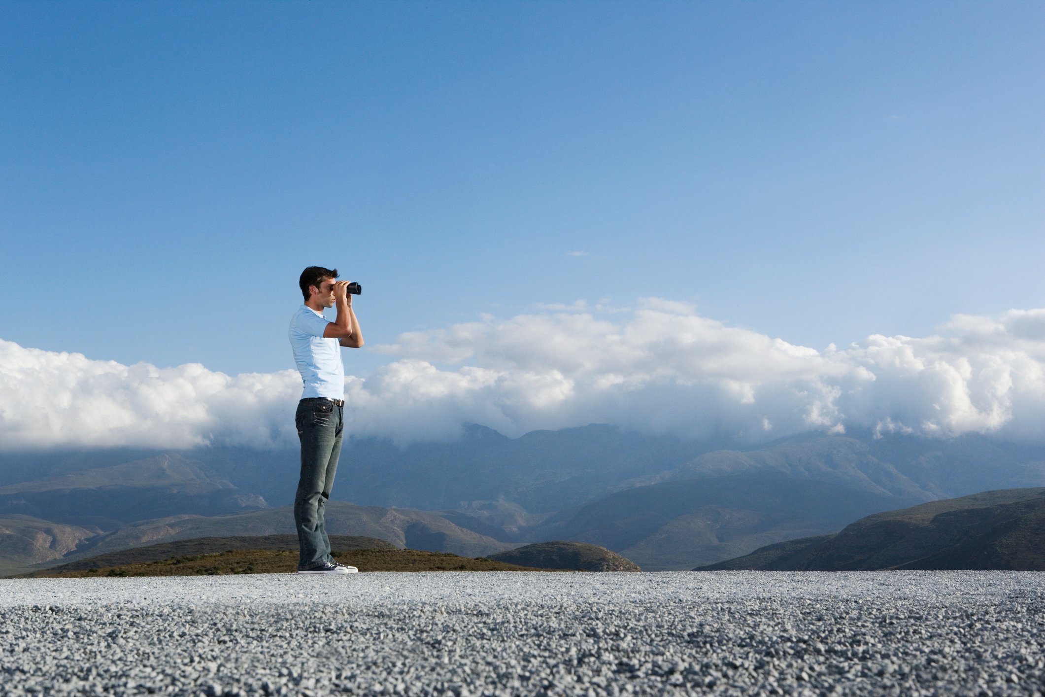 A man looks through binoculars, with a blue sky and mountains behind him.
