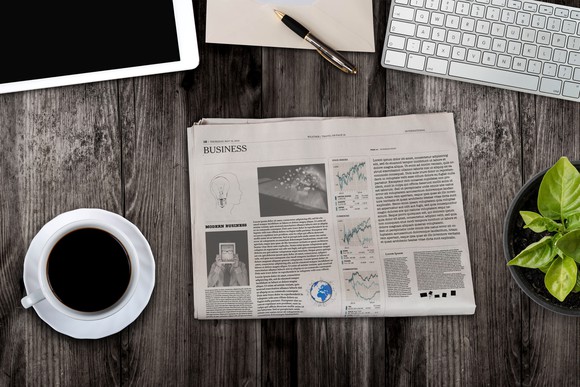 Newspaper on a table next to a cup of coffee, a tablet, a keyboard, and a notebook