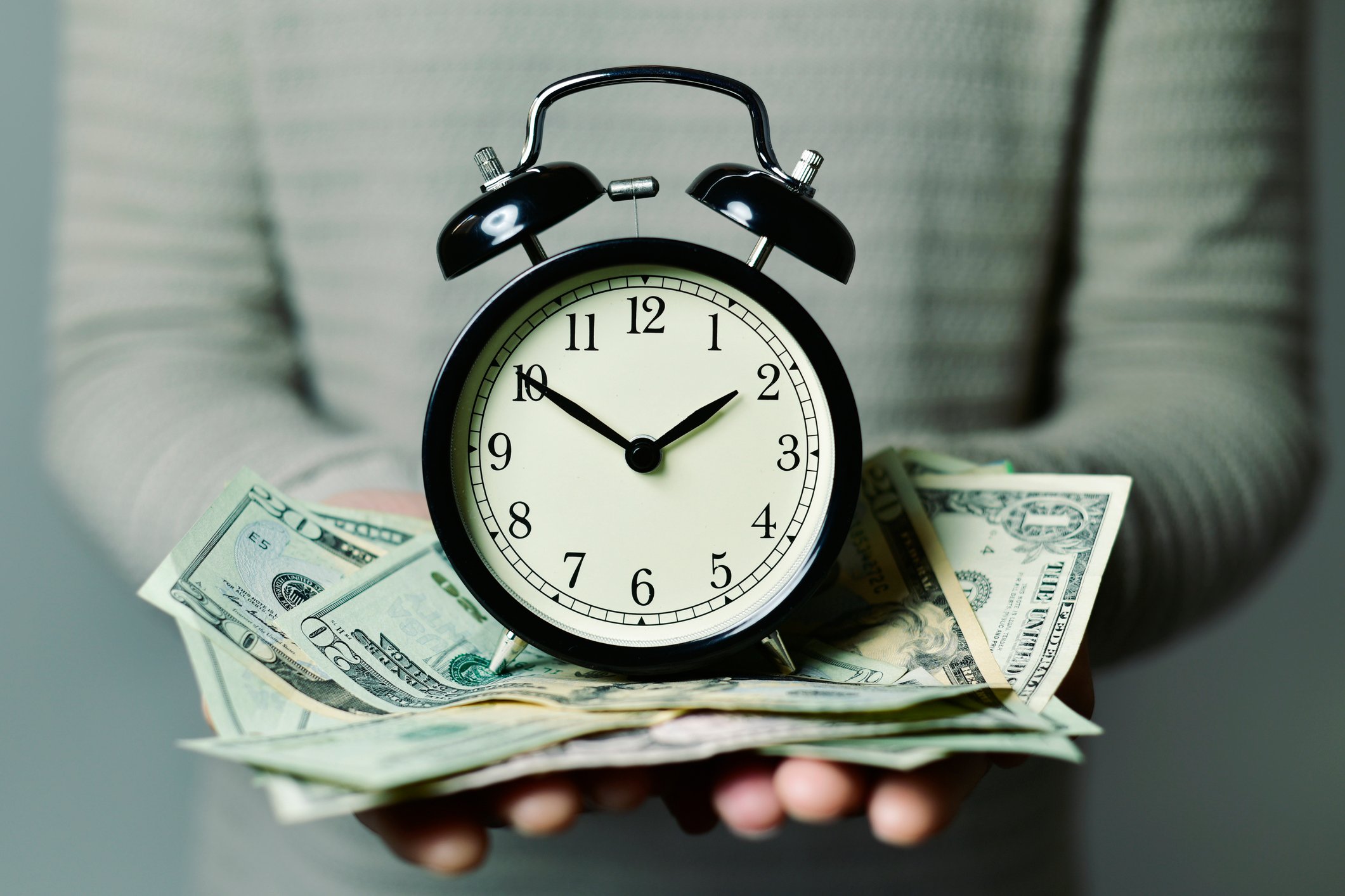 An old fashioned clock sits on top of a pile of money in someone's hands.