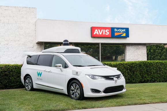 A Waymo vehicle in front of a building bearing the Avis/Budget logo.