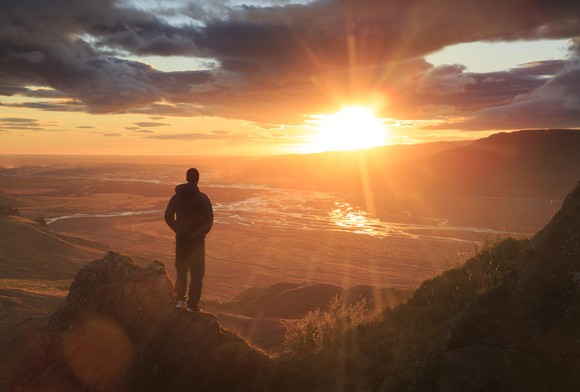From a mountaintop, a man looks out at the sun on the horizon. 