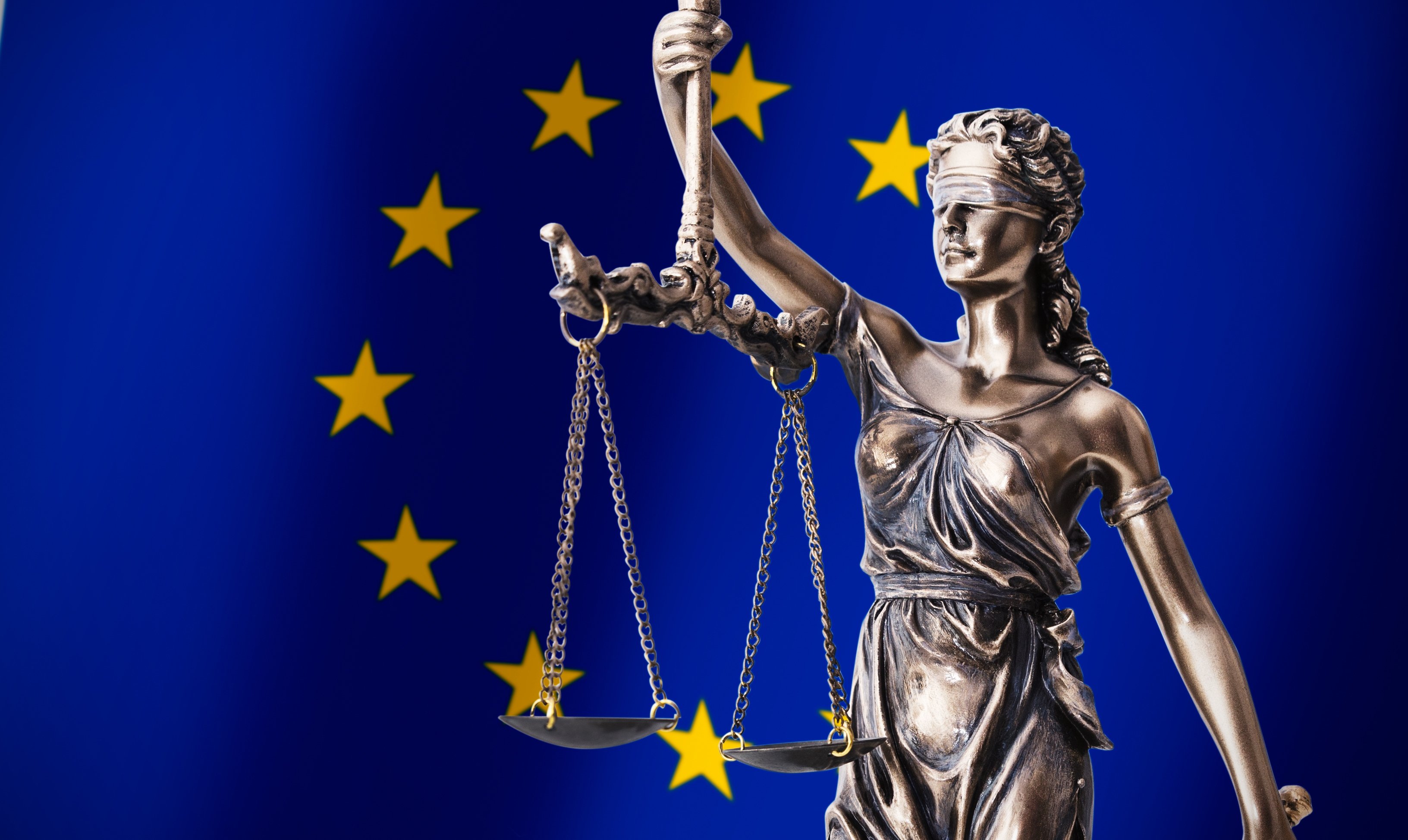 Lady Justice statue (blindfolded woman holding balance scales) with the flag of Europe as a backdrop.