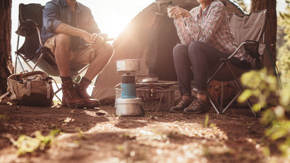 Two campers sit in the woods, surrounded by their camping gear.