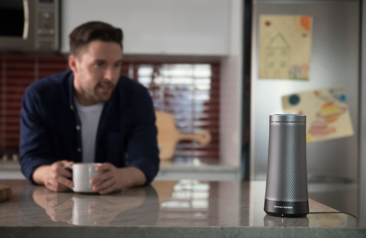 Cylindrical smart speaker on kitchen counter, while man with cup of coffee speaks to it.