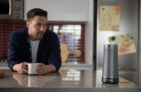 Cylindrical smart speaker on kitchen counter, while man with cup of coffee speaks to it.