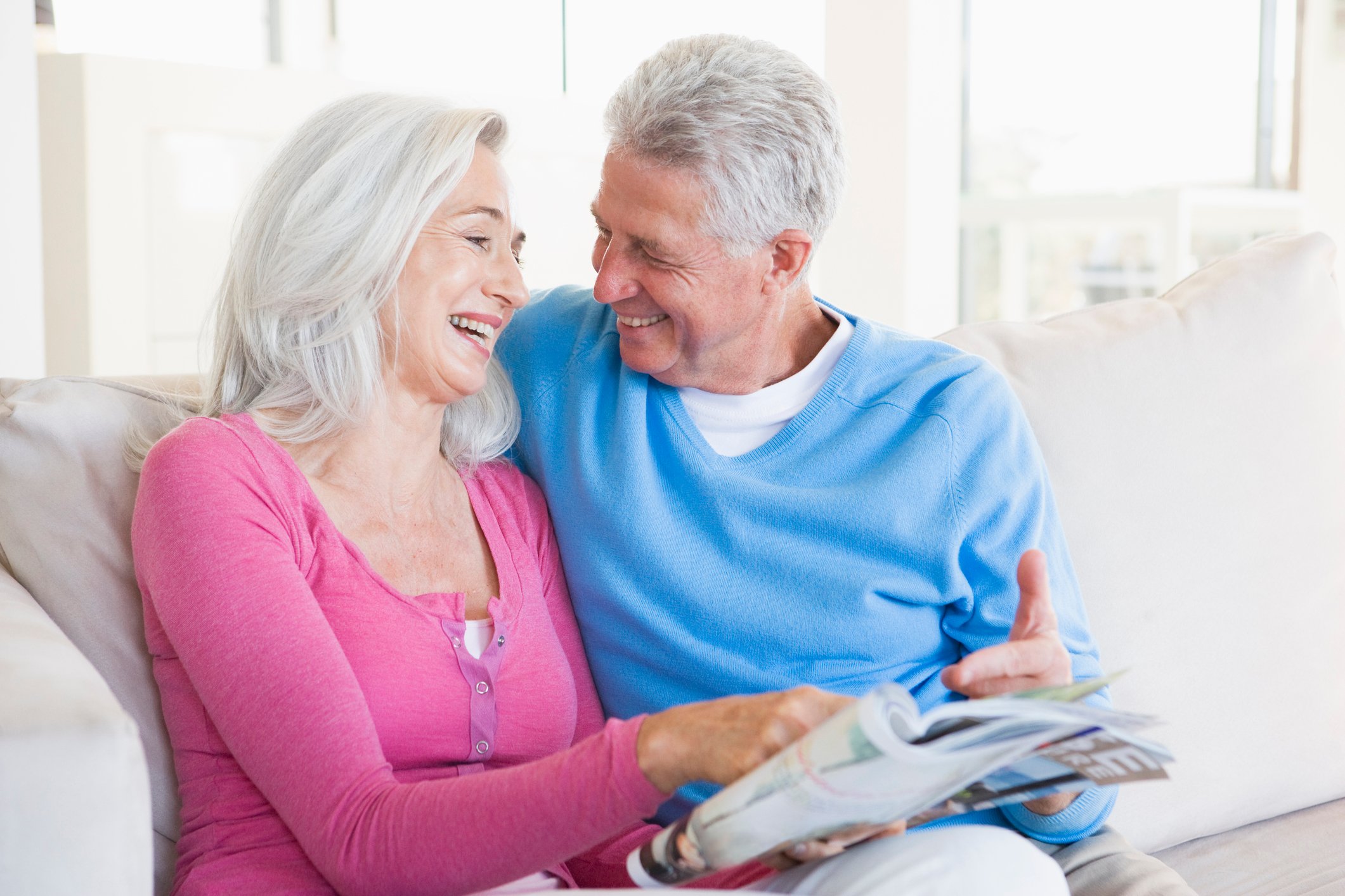 A retired couple sits smiling side by side on a sofa.