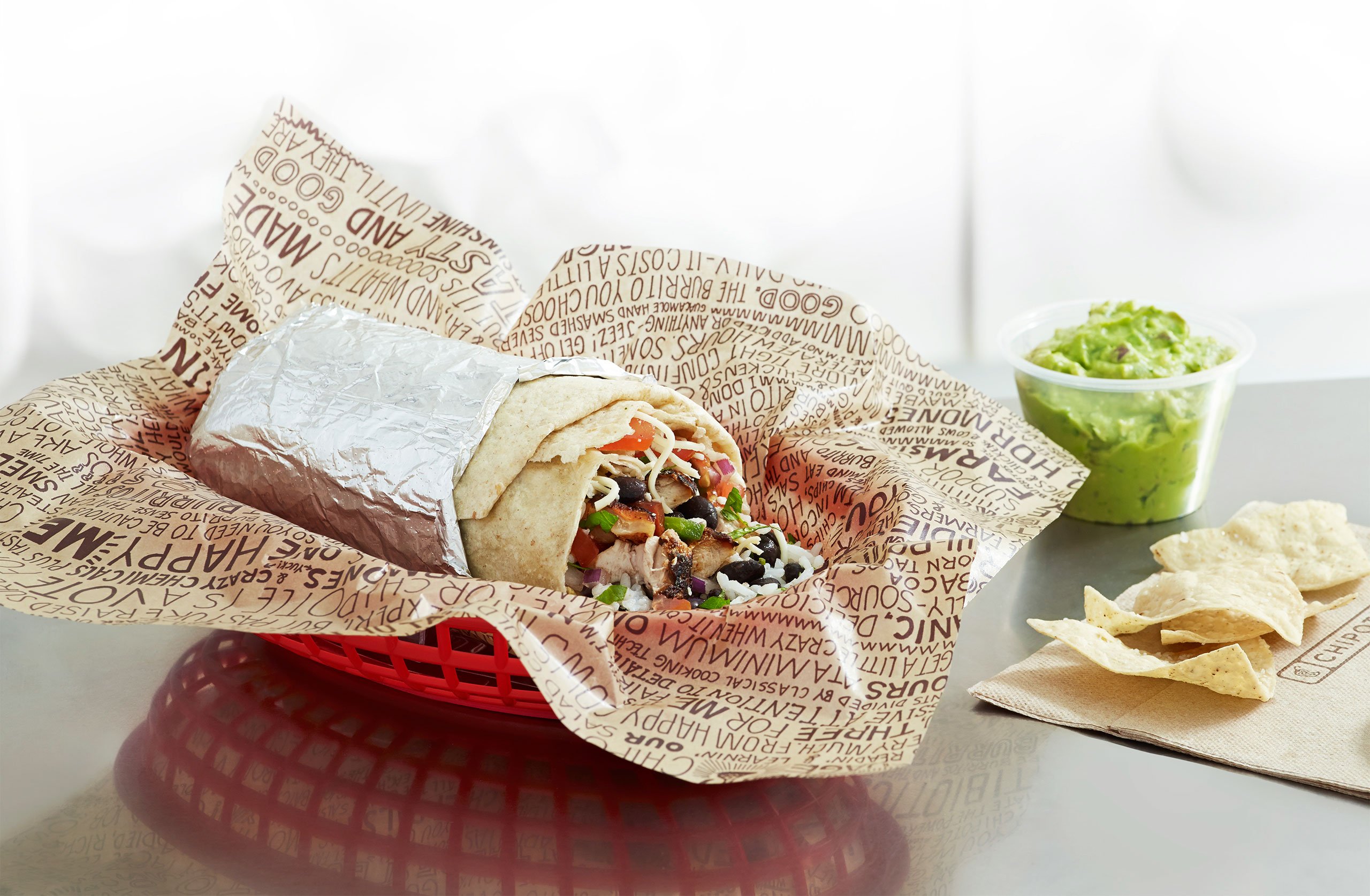 Chipotle burrito wrapped, laying in a red basket, with chips and guacamole on the side.