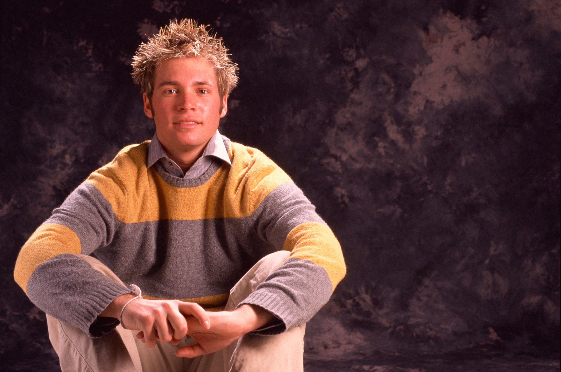 A crouched young man in a pullover displays preppy fashion style.