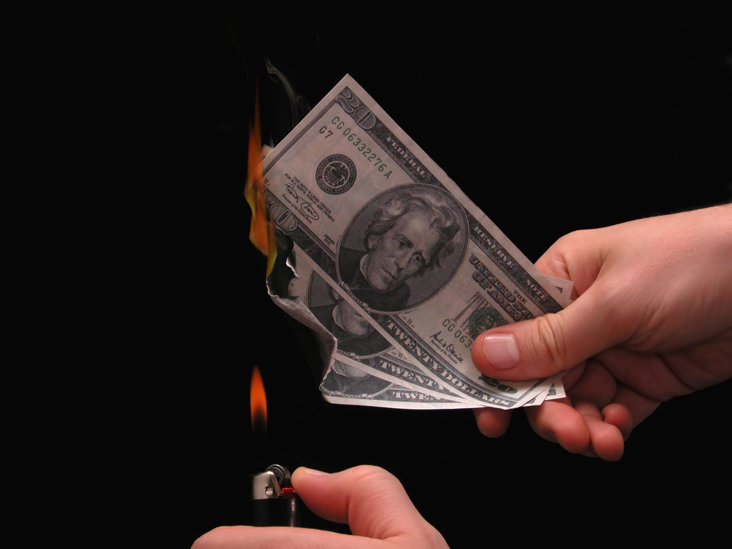 Hands holding a lighter and some burning 20-dollar bills on a black background.