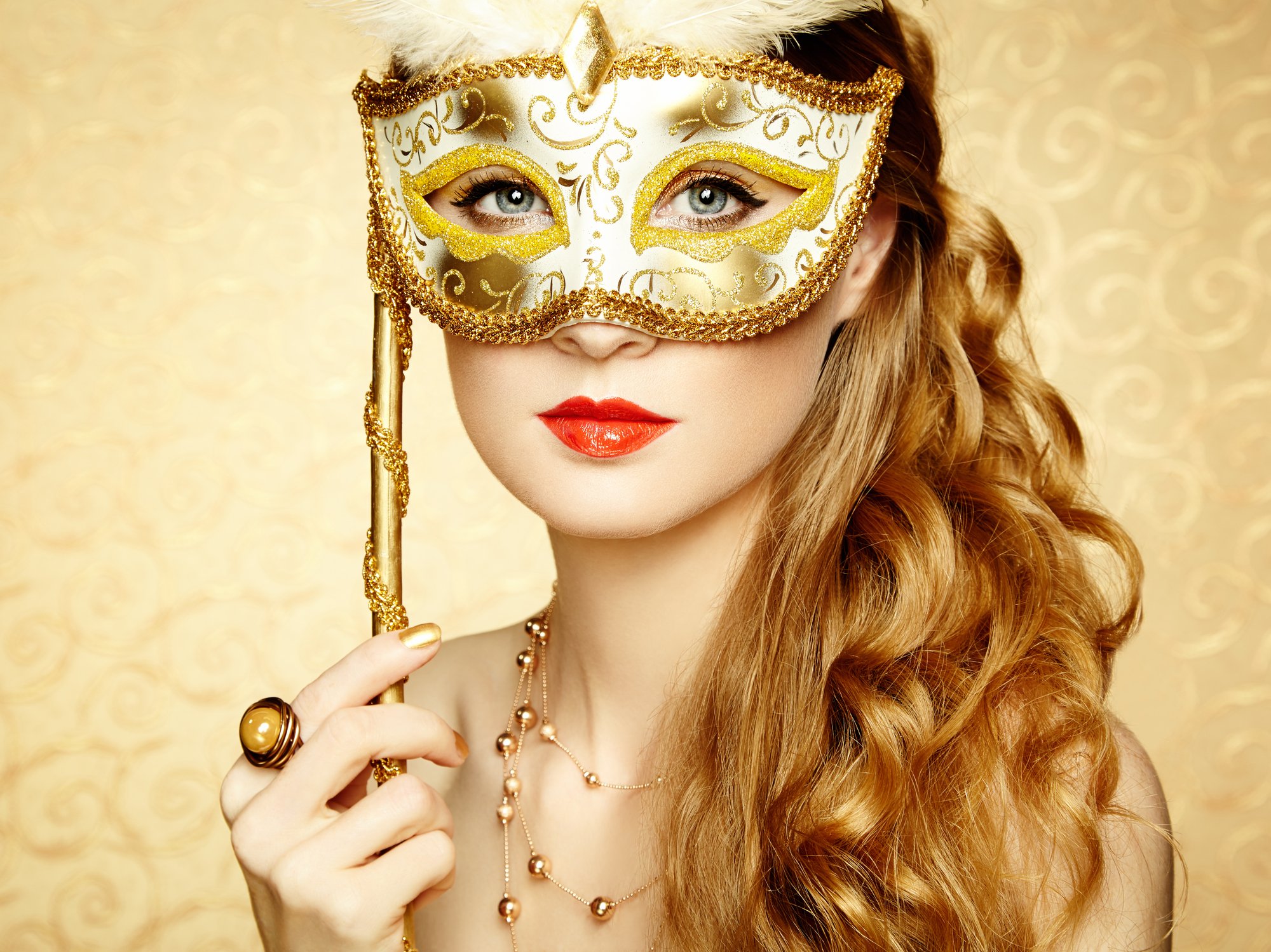 A woman holds a gold masquerade mask in front of her face.