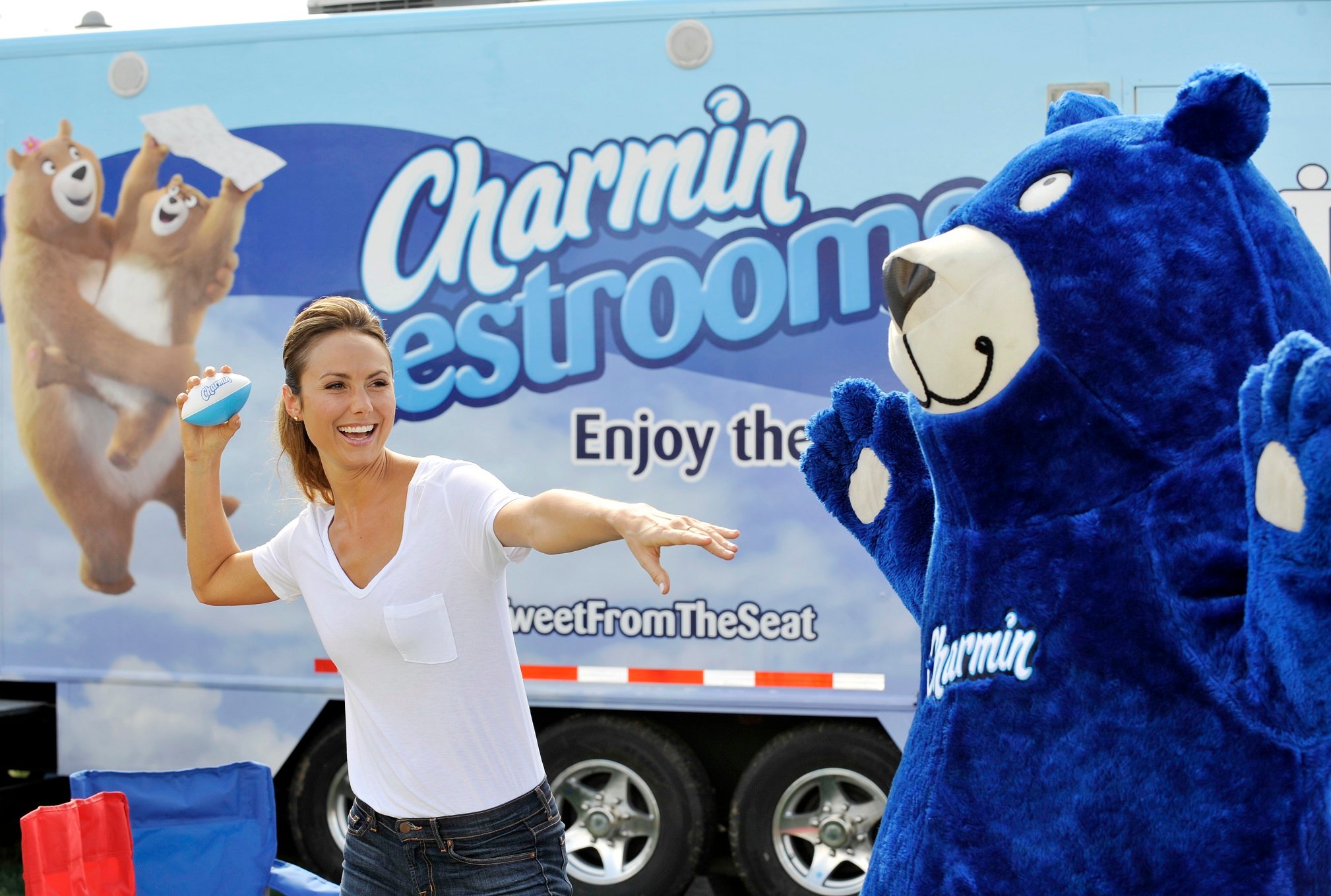 A person in a blue bear suit and a woman preparing to throw a ball both stand in front of a Charmin truck, in a marketing event.