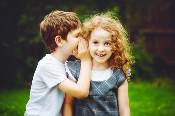 A small boy whispering in the ear of a young girl.