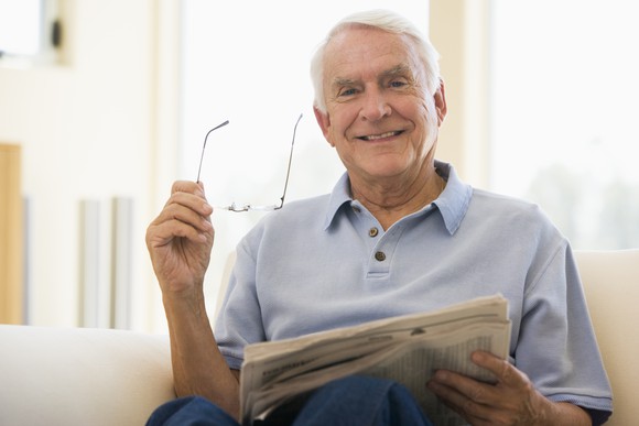 A senior man sits on a sofa and smiles, holding a newspaper in one hand and reading glasses in another.