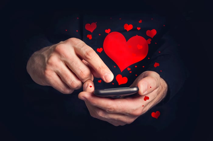 Two hands, one holding a cellphone, one swiping the screen from which red hearts are floating.