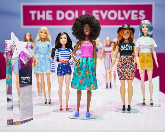 A lineup of diverse dolls standing in front of a banner reading "The Doll Evolves" with the Doll of the Year Award.