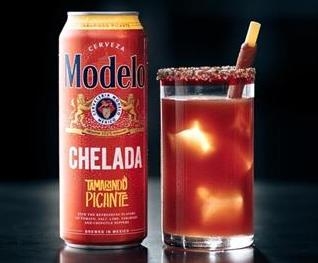 Festive can of Modelo Chelada "Tamorindo Picante" next to a glass filled with the reddish orange beverage, black background.