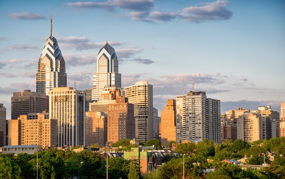 The Philadelphia skyline, with green trees in the foreground and a partly cloudy sky in the background.