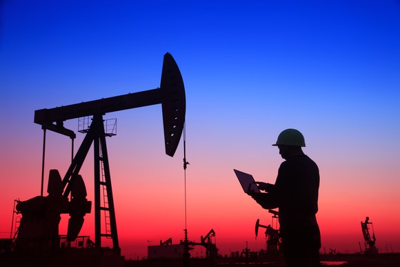 An oil-field worker with a laptop stands beneath a pumpjack at sunset.
