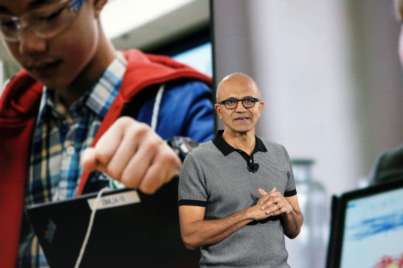 Satya NAdella speaking on a stage with picture of young boy operating a piece of machinery behind him. 