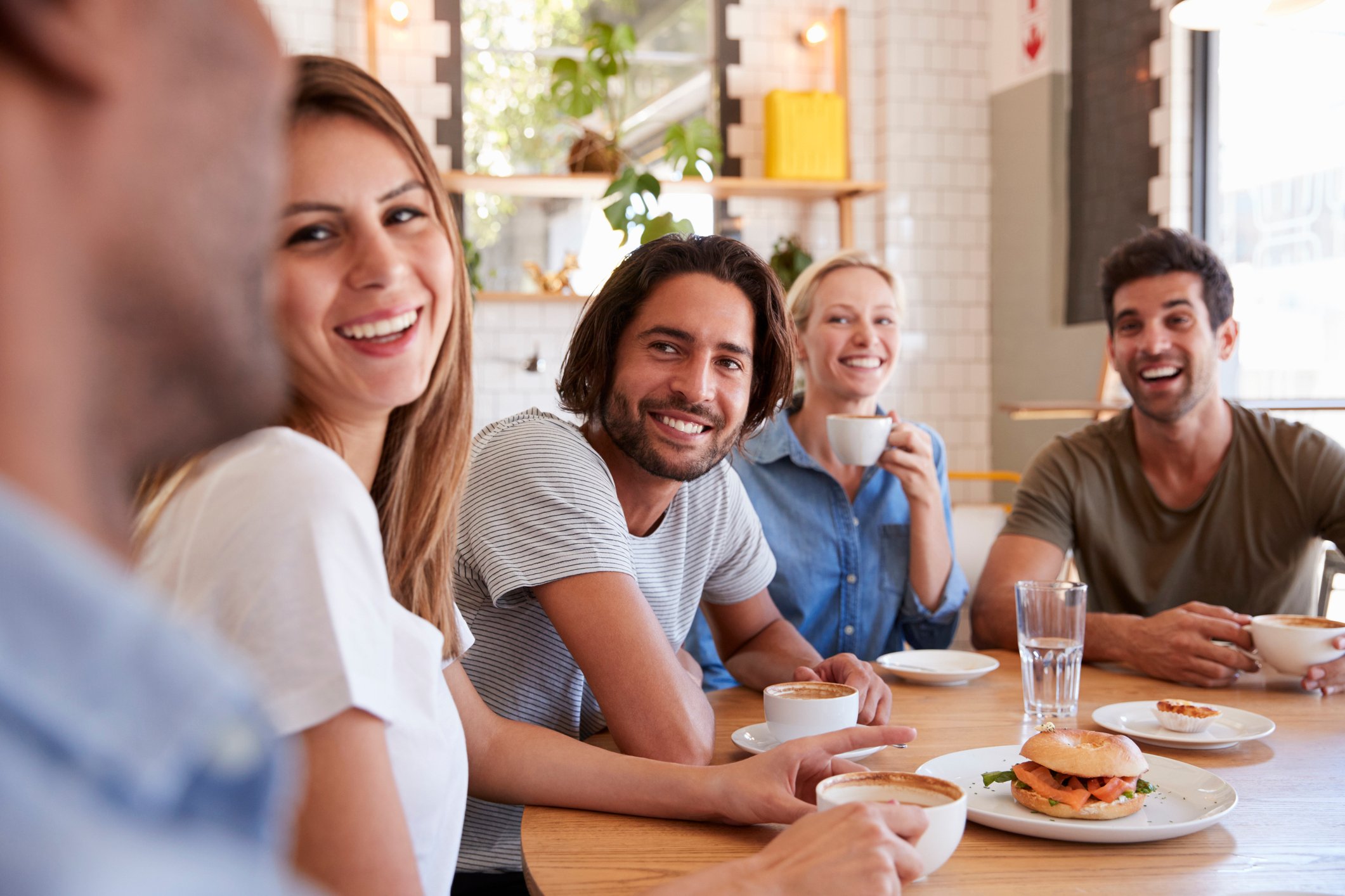 A group of millennials smile as they sit around a table, with food and drink in front of them. 