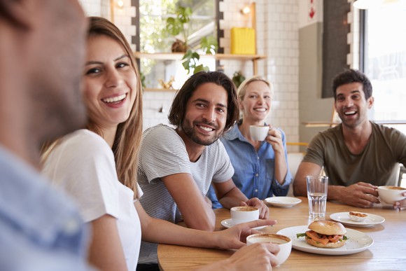 A group of millennials smile as they sit around a table, with food and drink in front of them. 