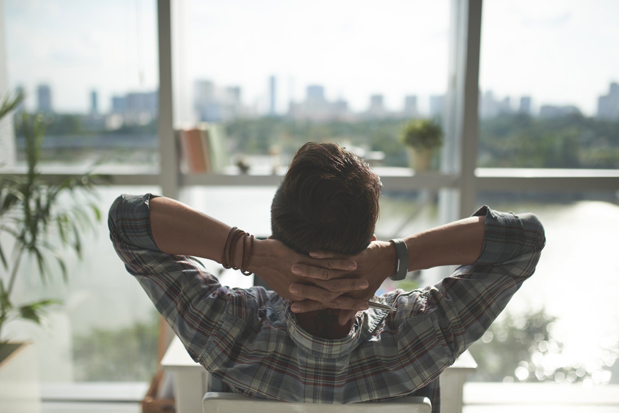 A man leans back and relaxes in a chair with his hands behind his head, as he looks out at a city skyline.