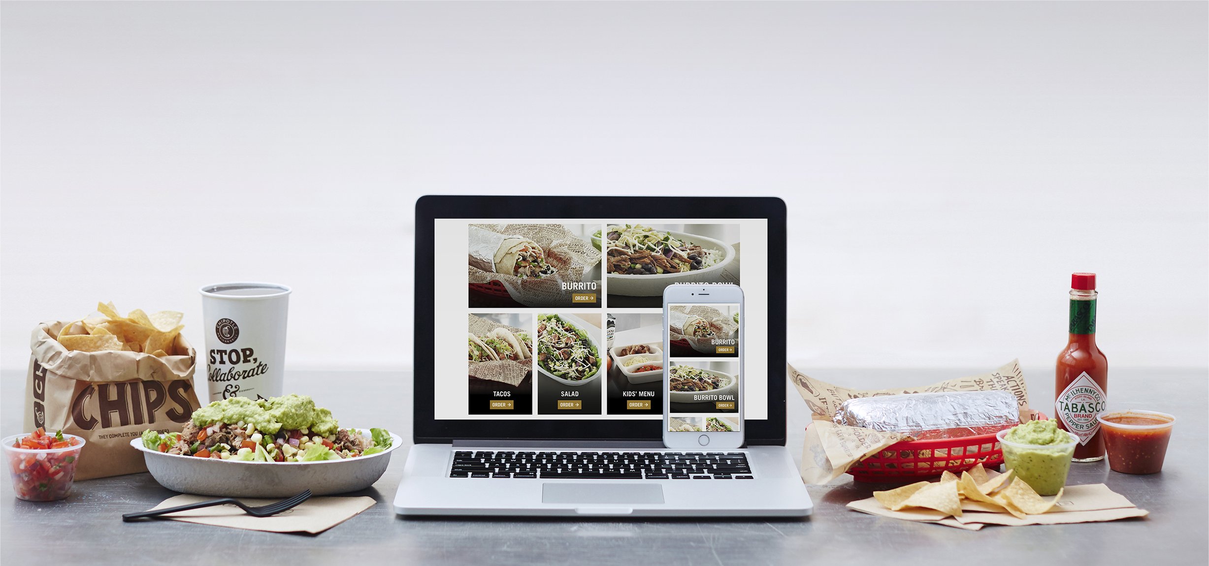 An assortment of Chipotle menu items are spread out on a tabletop. Between them sit a laptop and smartphone showing Chipotle's app.