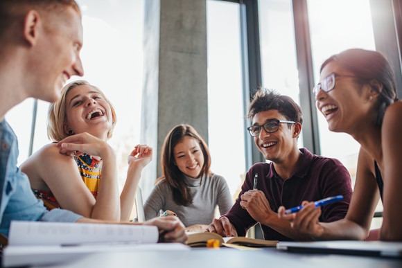 College-age men and women laughing together around a table with open books on it.