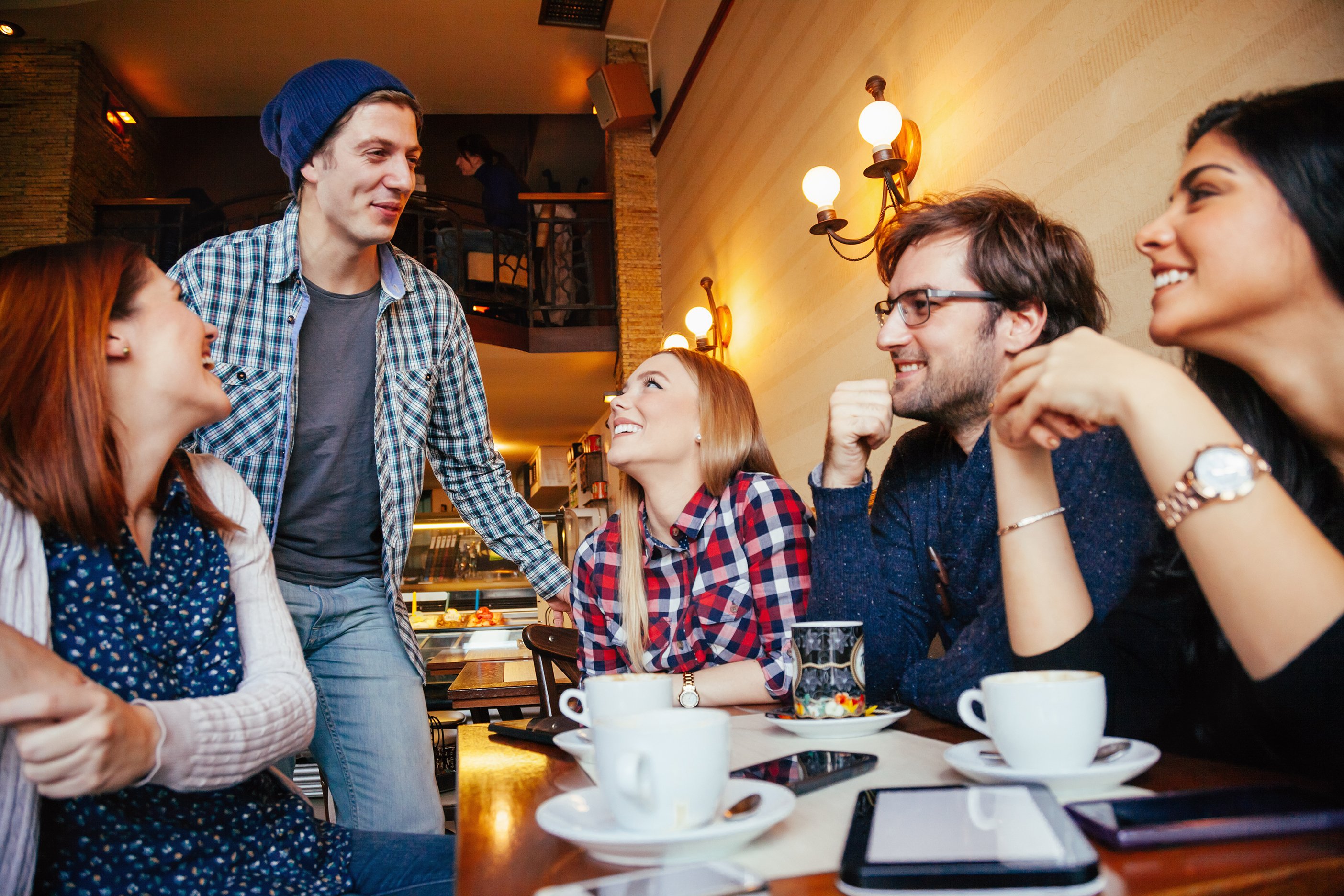 A group of young people, smiling and gathered around a cafe table, with coffee cups and a tablet on the table in front of them. 