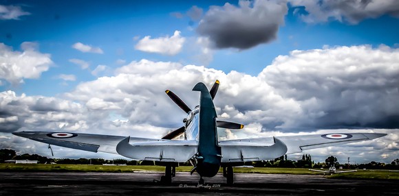 A British Spitfire, a World War II fighter plane, sits on a runway against blue sky with clouds.