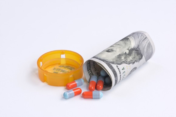 Capsules, colored red and blue, spill out of a hundred-dollar bill wrapped up to look like a tipped-over prescription bottle.